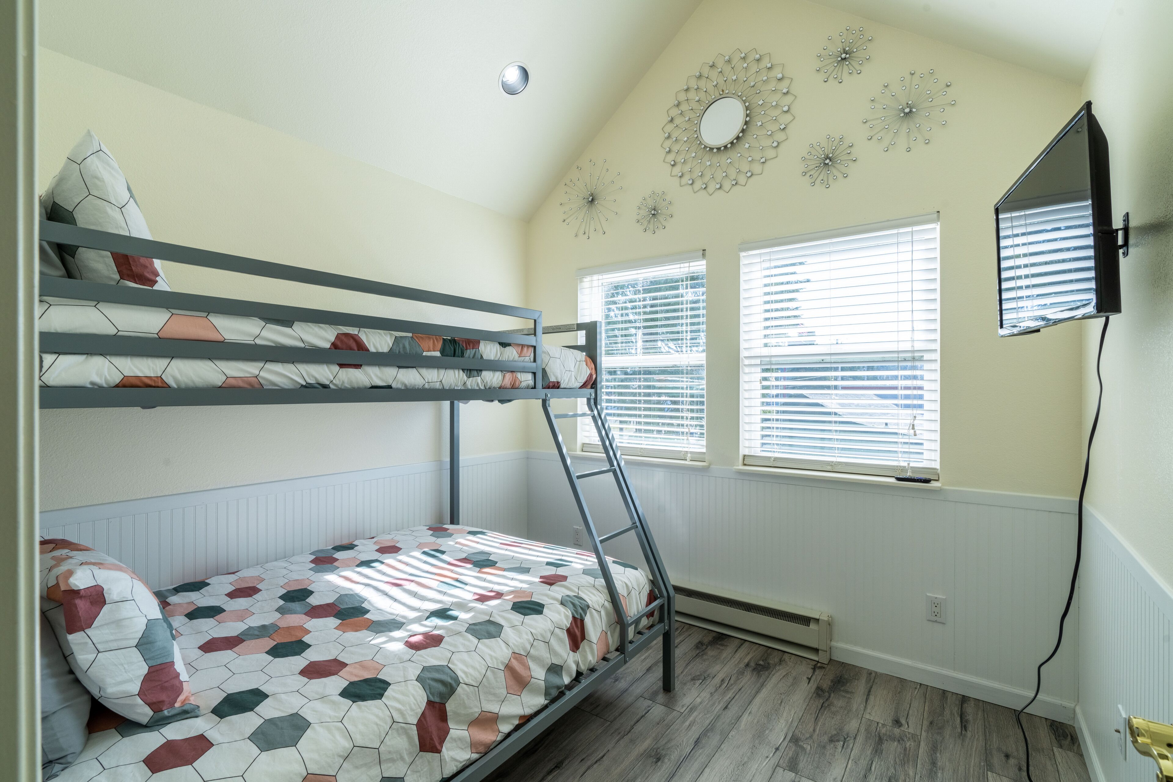 A bunk bed room with a TV mounted on the wall, a window with blinds, and decorative wall art.