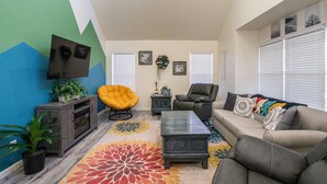 Uma sala de estar com sofá, cadeiras, mesa de centro, televisão e tapete colorido.