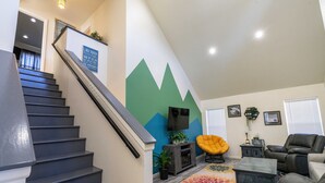 Uma escadaria leva a uma área de estar com um mural verde na parede, uma TV de tela plana, uma poltrona amarela e um sofá cinza.