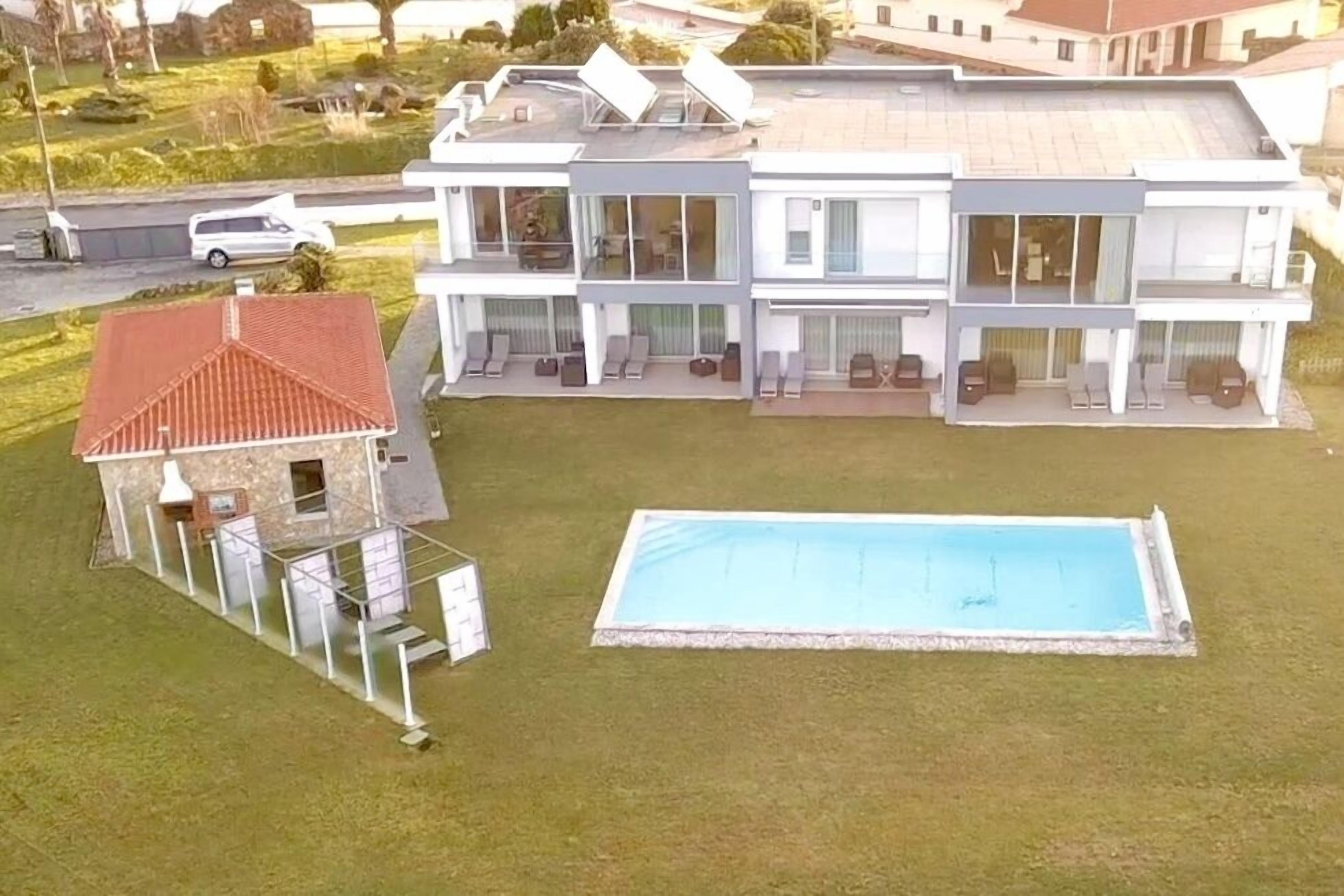 A modern two-story house with a swimming pool, a separate building with a red roof, and a street in the background.
