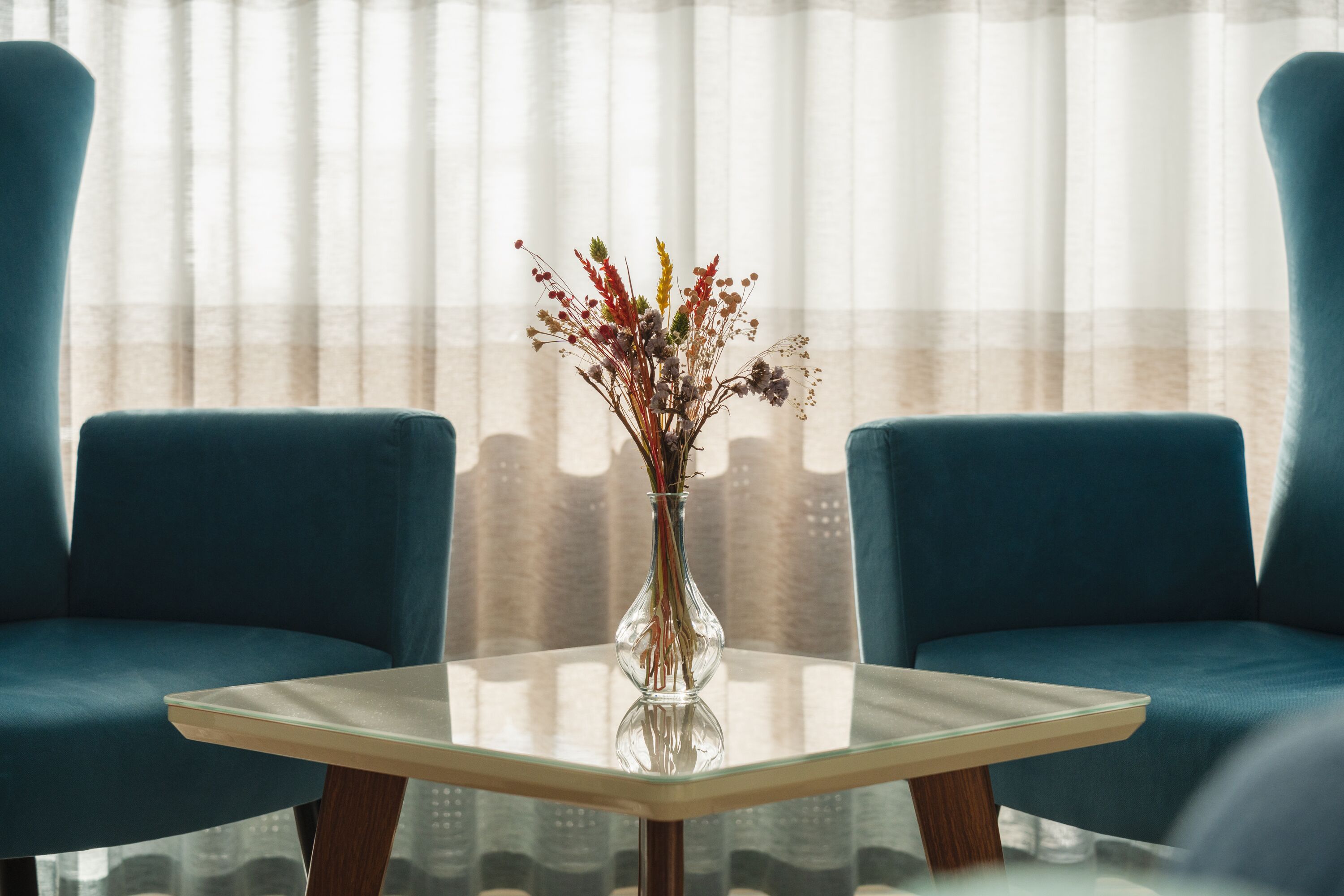Een kamer met een glazen tafel, twee blauwe stoelen en een vaas met gedroogde bloemen.