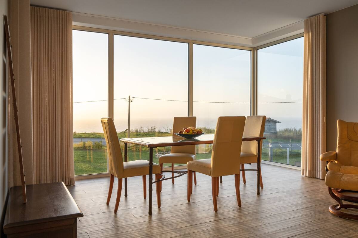 Un comedor con mesa y sillas, ventanas amplias que dan a un paisaje, y piso de madera.