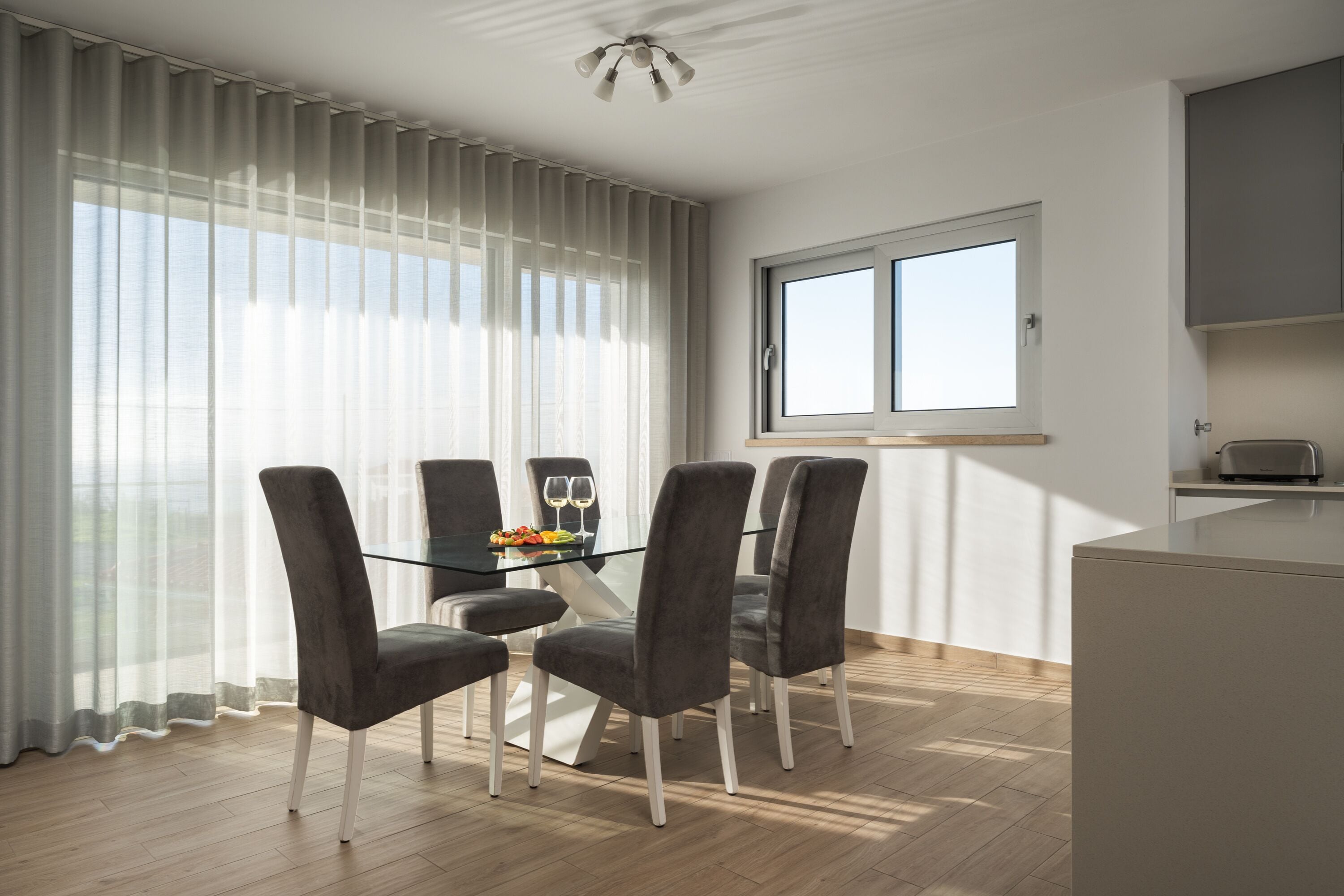 Un comedor moderno con una mesa de vidrio, cuatro sillas grises tapizadas y vistas al exterior a través de cortinas translúcidas.