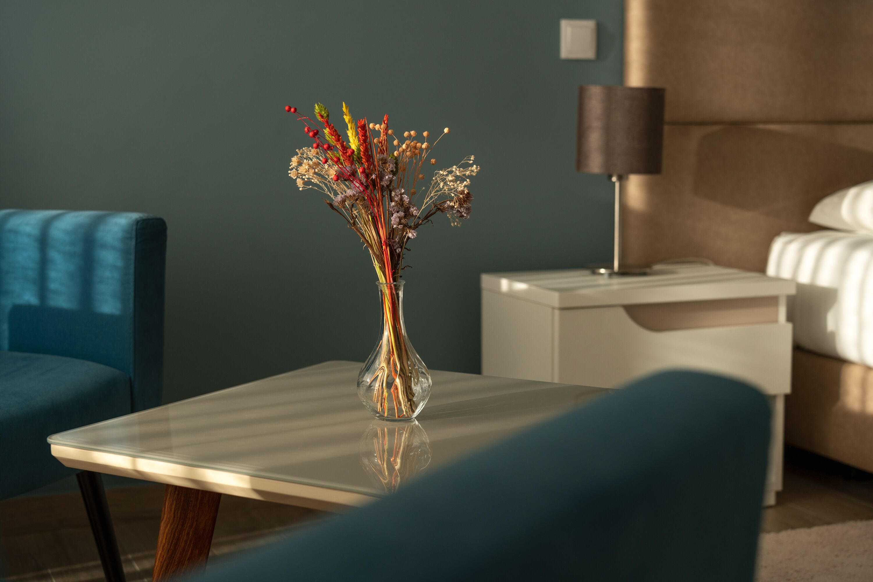 A modern hotel room with a glass table, a vase with flowers, a blue armchair, a white bedside table, and a lamp.