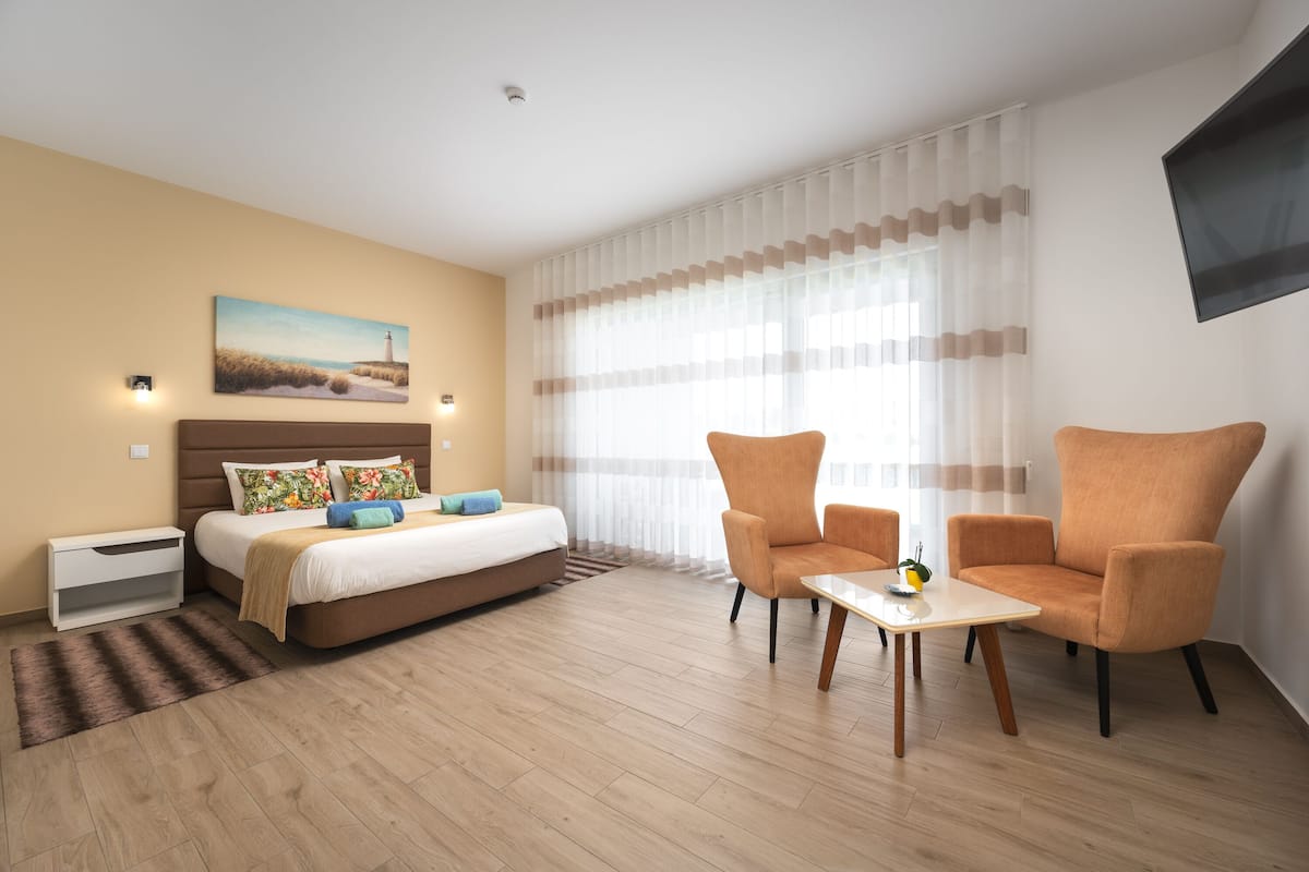 Ett modernt hotellrum med en säng, ett skrivbord, två fåtöljer, en tavla och ett stort fönster med tunna, genomskinliga gardiner.