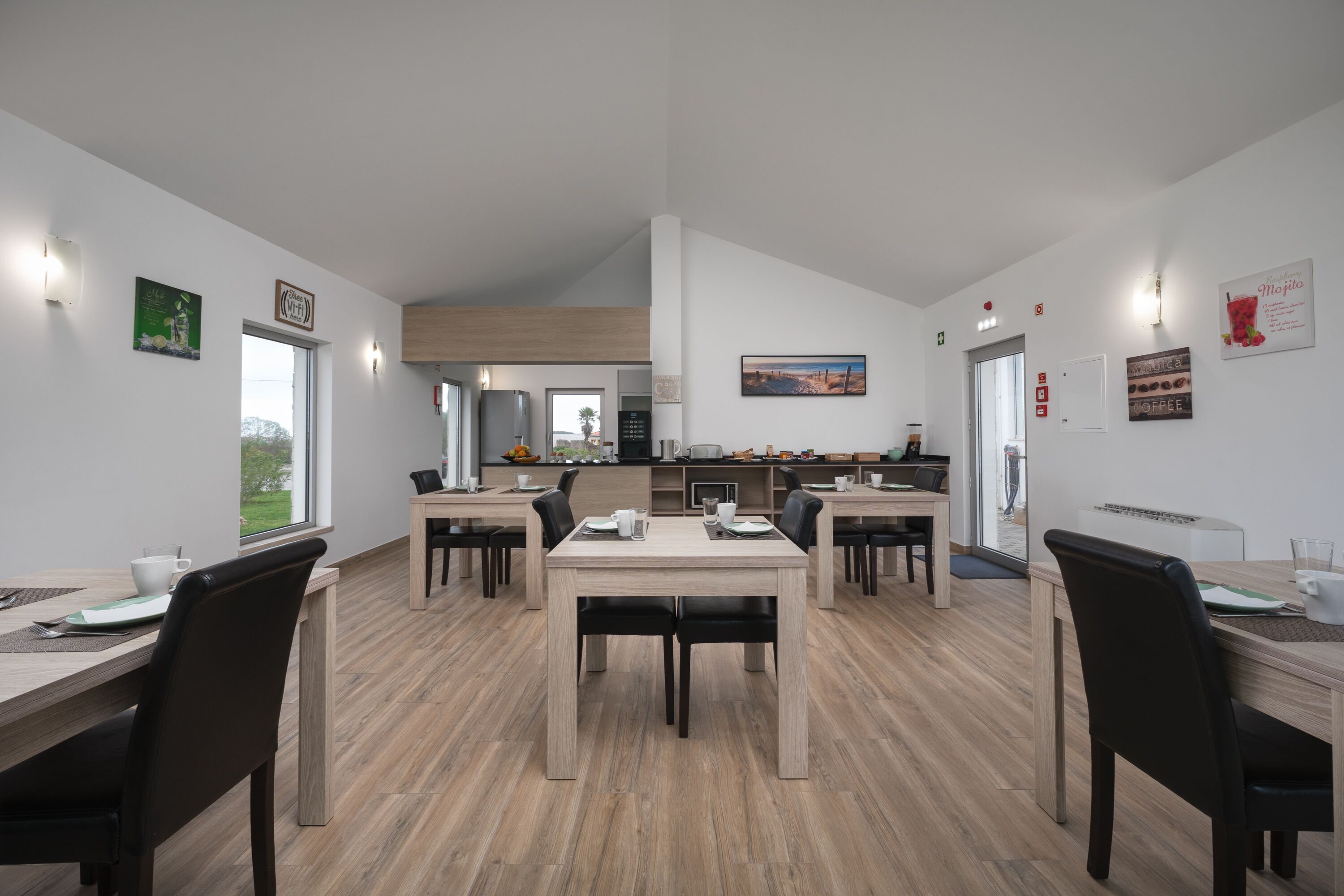 A modern dining area with wooden tables and chairs, a kitchen in the background, and a view of the outdoors.