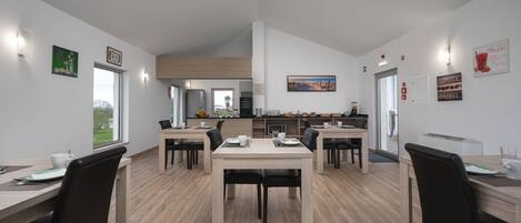 Un espace repas moderne avec des tables et des chaises en bois, une cuisine en arrière-plan et une vue sur l’extérieur.