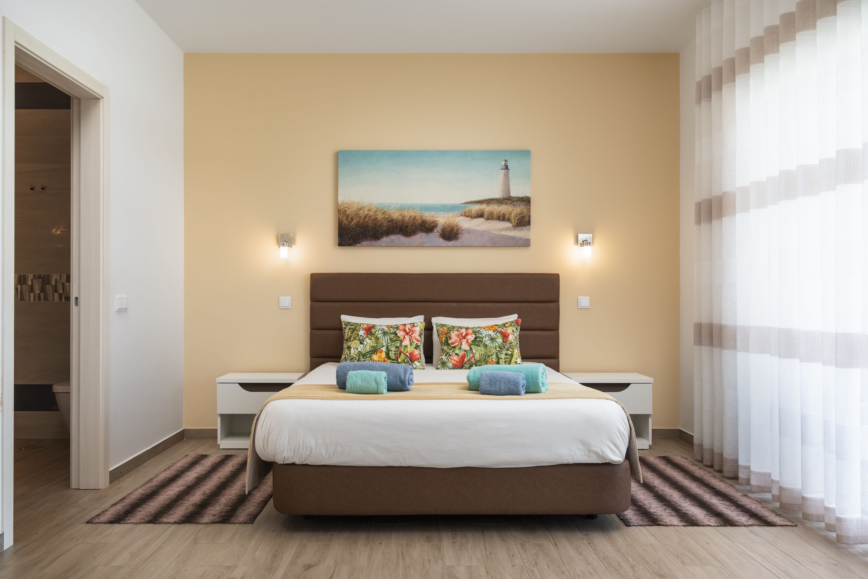 A bedroom with a large bed, two pillows, a nightstand, a painting on the wall, and a bathroom visible through an open door.