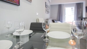 Una mesa de comedor dispuesta con platos blancos, cubiertos y copas de vino, con un sofá y una ventana al fondo.