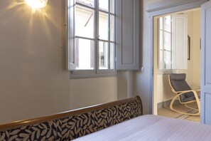 Une chambre à coucher avec un lit, une fenêtre avec des volets, une chaise et une lampe fixée au mur.