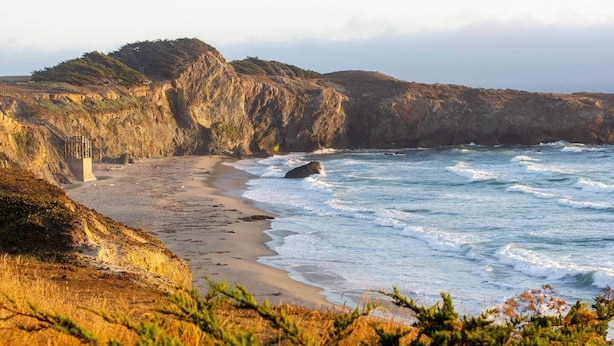 A coastal landscape with a rocky cliff, a sandy beach, and a staircase leading down to the beach.