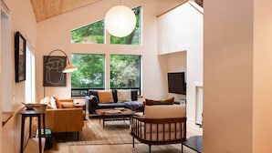 A modern living room with a sofa, armchair, and coffee table, large windows with a view of greenery, and a wooden floor.