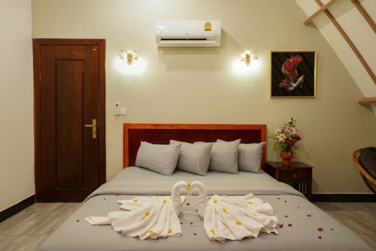 A hotel room with a bed, a door, an air conditioning unit, a wall-mounted light fixture, a painting, and a small table with a flower vase.