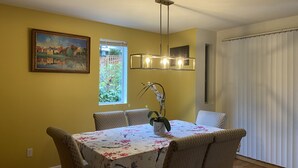 Uma sala de jantar com uma mesa posta para quatro pessoas, rodeada por cadeiras. Há uma janela com vista para o verde lá fora, um quadro na parede e um lustre sobre a mesa.