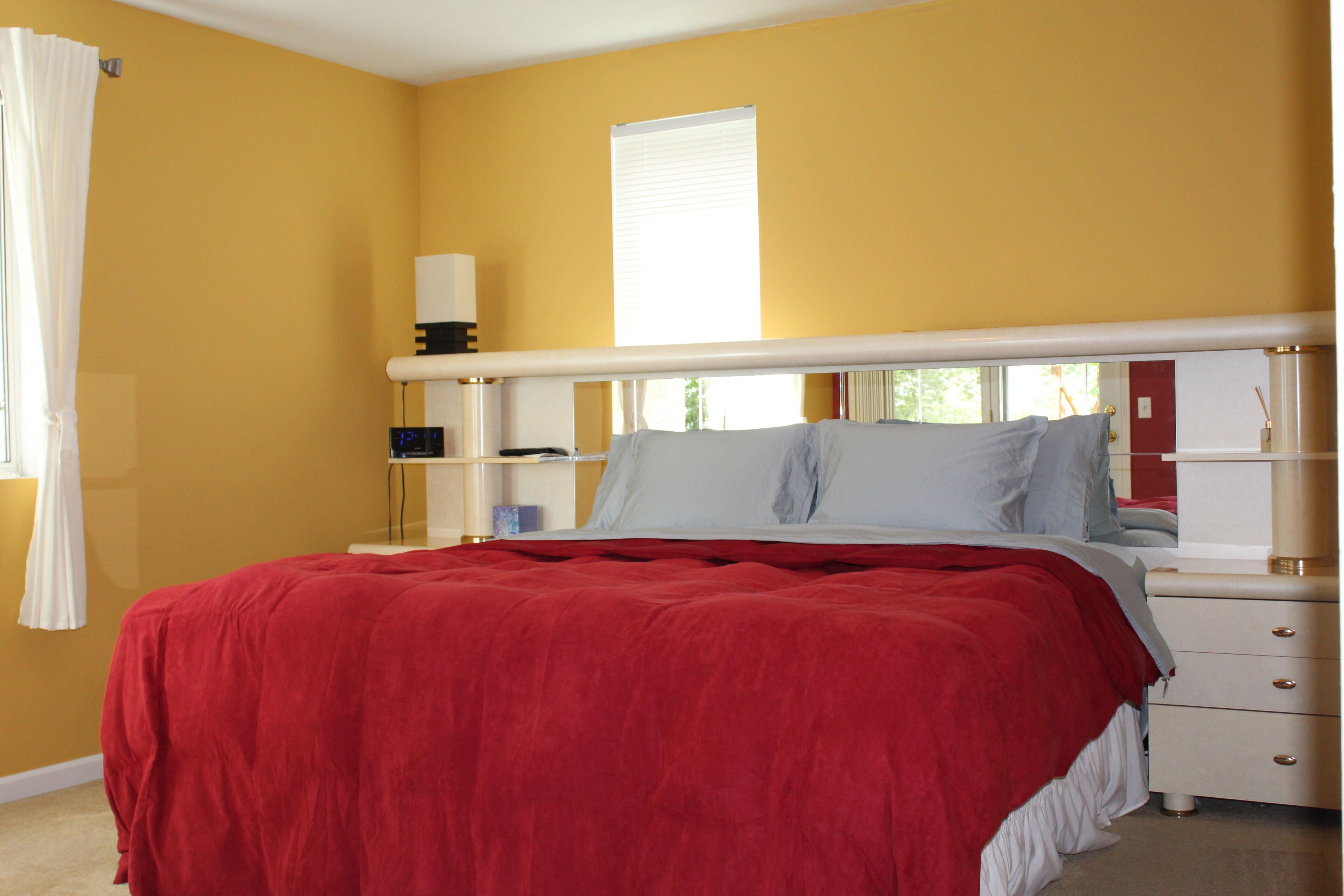A bedroom with a red bedspread, a white headboard, a bedside table with a lamp, and a window with blinds.
