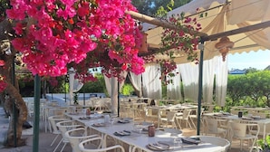 An outdoor dining area with white tables and chairs, covered by a canopy, and surrounded by greenery. - Villa with outdoor spaces (Cabanas de tavira)