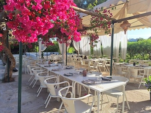 An outdoor dining area with white tables and chairs, covered by a canopy, and surrounded by greenery.