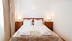 Una cama doble con dos almohadas blancas y una manta a juego, flanqueada por mesitas de noche de madera con lámparas, en una habitación con colcha de estampado floral y una lámpara de pared.