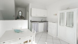 A small, white kitchen with a tiled floor, a white table with a blue object on it, and white cabinets.