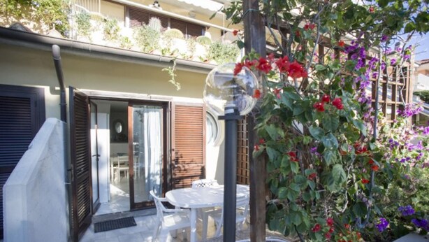 A patio with a table and chairs, a glass door leading to an interior space, and a garden with various plants and flowers.