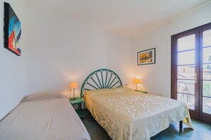 A bedroom with two single beds, a green metal headboard, a small table, two bedside lamps, and a window with a view of buildings.