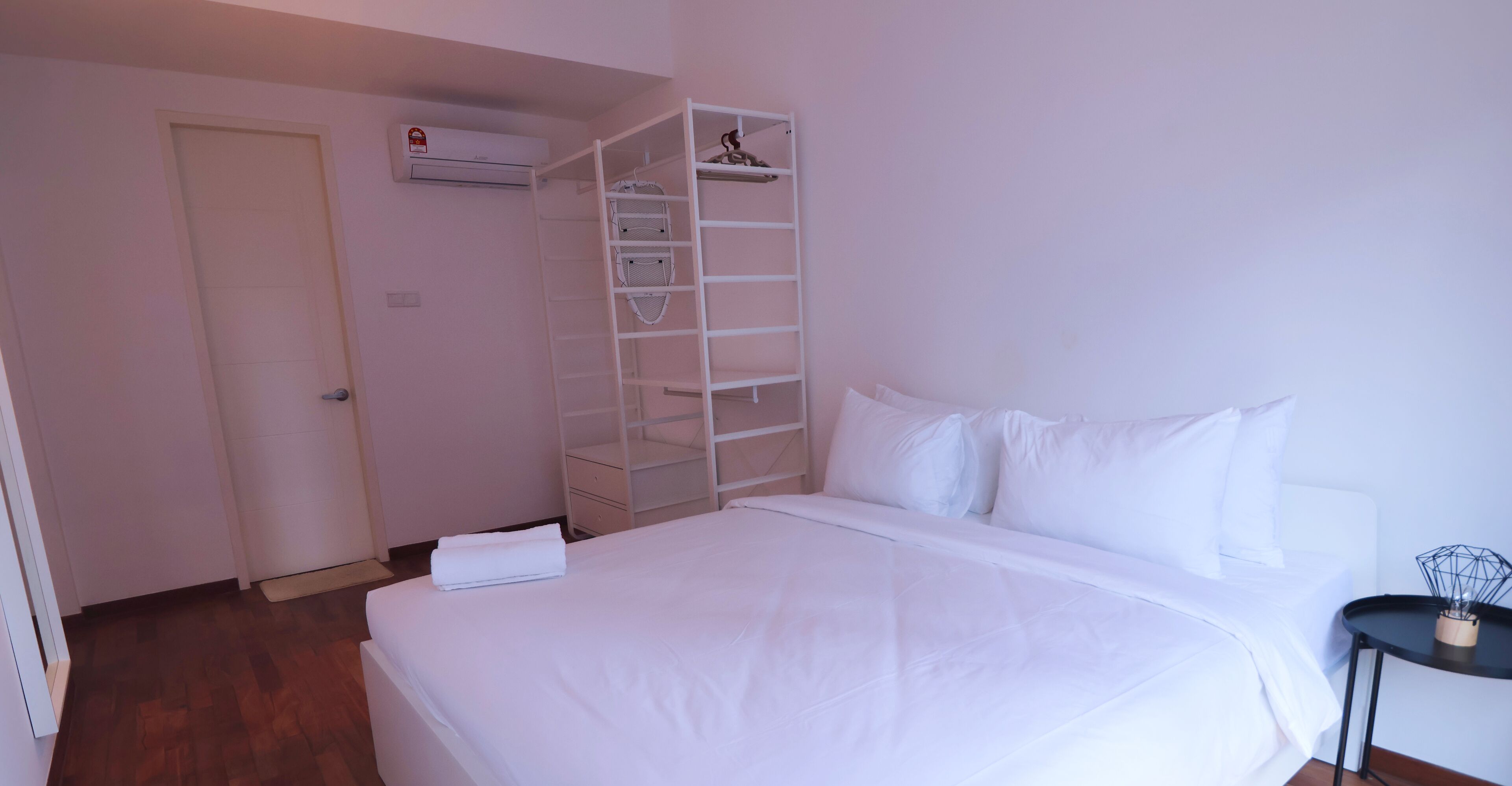 A bedroom with a neatly made bed, a white wardrobe with shelves, a small bedside table, and an air conditioning unit on the wall.