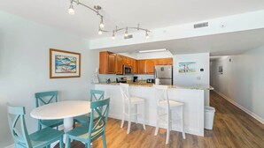 A modern kitchen with wooden cabinets, a white island with bar stools, and a dining area with teal chairs. - Gulf-view condo in Destin with pool, hot tub and beach access (Destin)