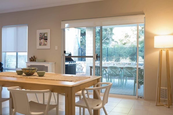 Uma sala de jantar com mesa de madeira e cadeiras brancas, uma cozinha com churrasqueira e uma porta de vidro deslizante que dá para um pátio externo.