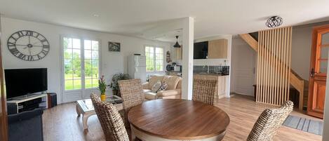A spacious living area with a round wooden dining table, wicker chairs, a sofa, and a television.