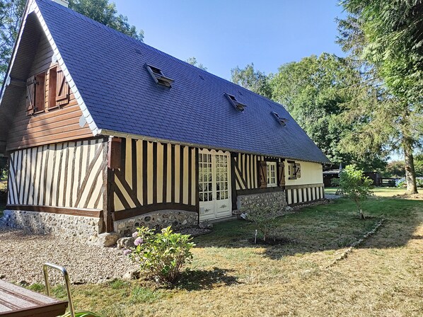 Une maison traditionnelle à colombages, dotée d’un toit à forte pente, de fondations en pierre et d’un petit jardin agrémenté d’un banc en bois et de fleurs.