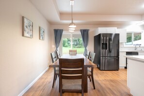 Una cocina moderna con un refrigerador de acero inoxidable, una mesa de comedor de madera y sillas. Hay una ventana con cortinas y vistas al exterior con vegetación.