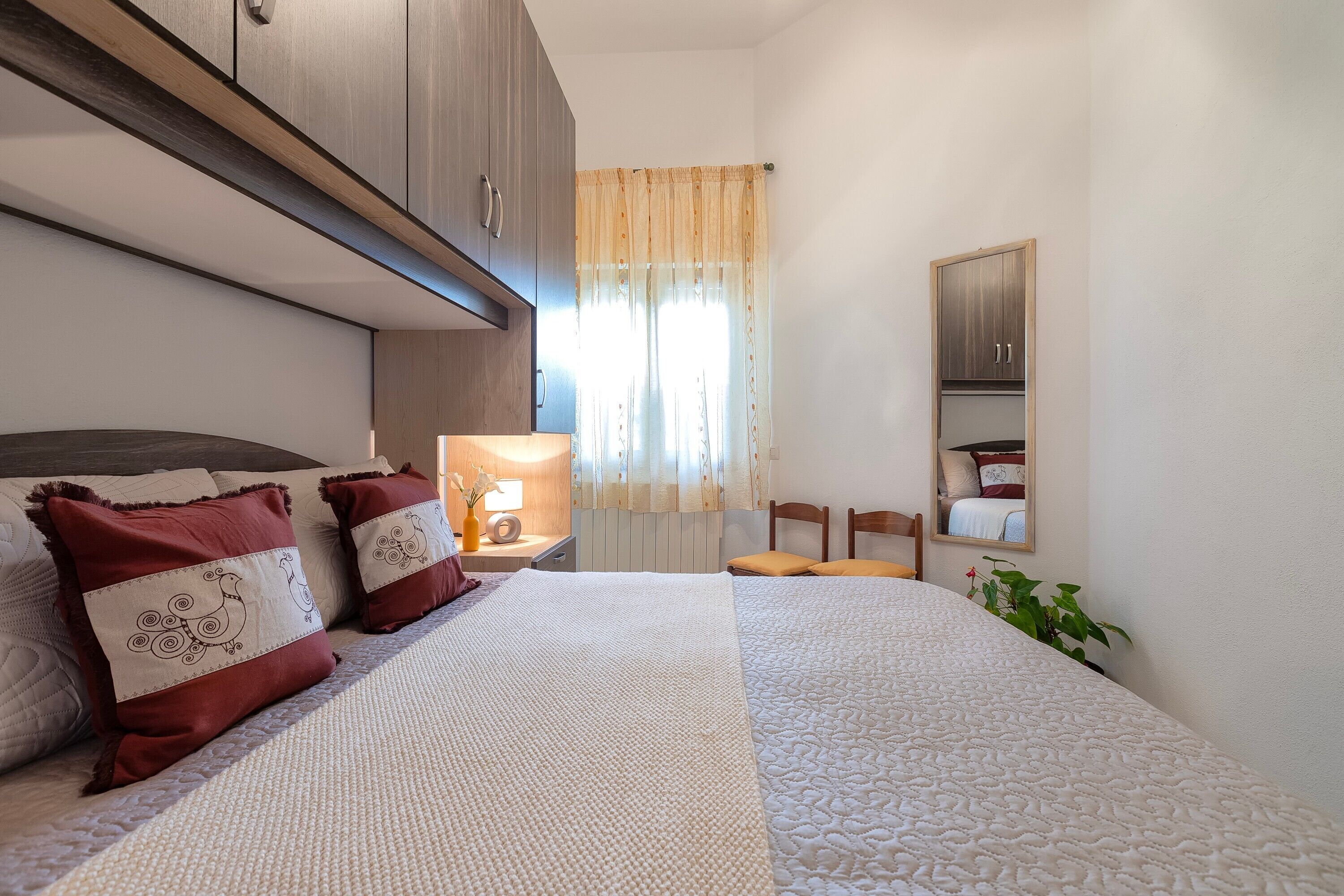 Un dormitorio con cama, mesita de noche, lámpara, espejo y planta en maceta.