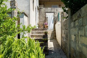 Un escalier étroit en pierre mène à une porte ornée d’un rideau, le tout étant entouré de verdure et d’un mur de pierre.