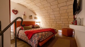 Ein Schlafzimmer mit schrägverdeckter Decke, einem Bett mit gemusterten Textilien, einer roten Bank und einem kleinen roten Hocker.