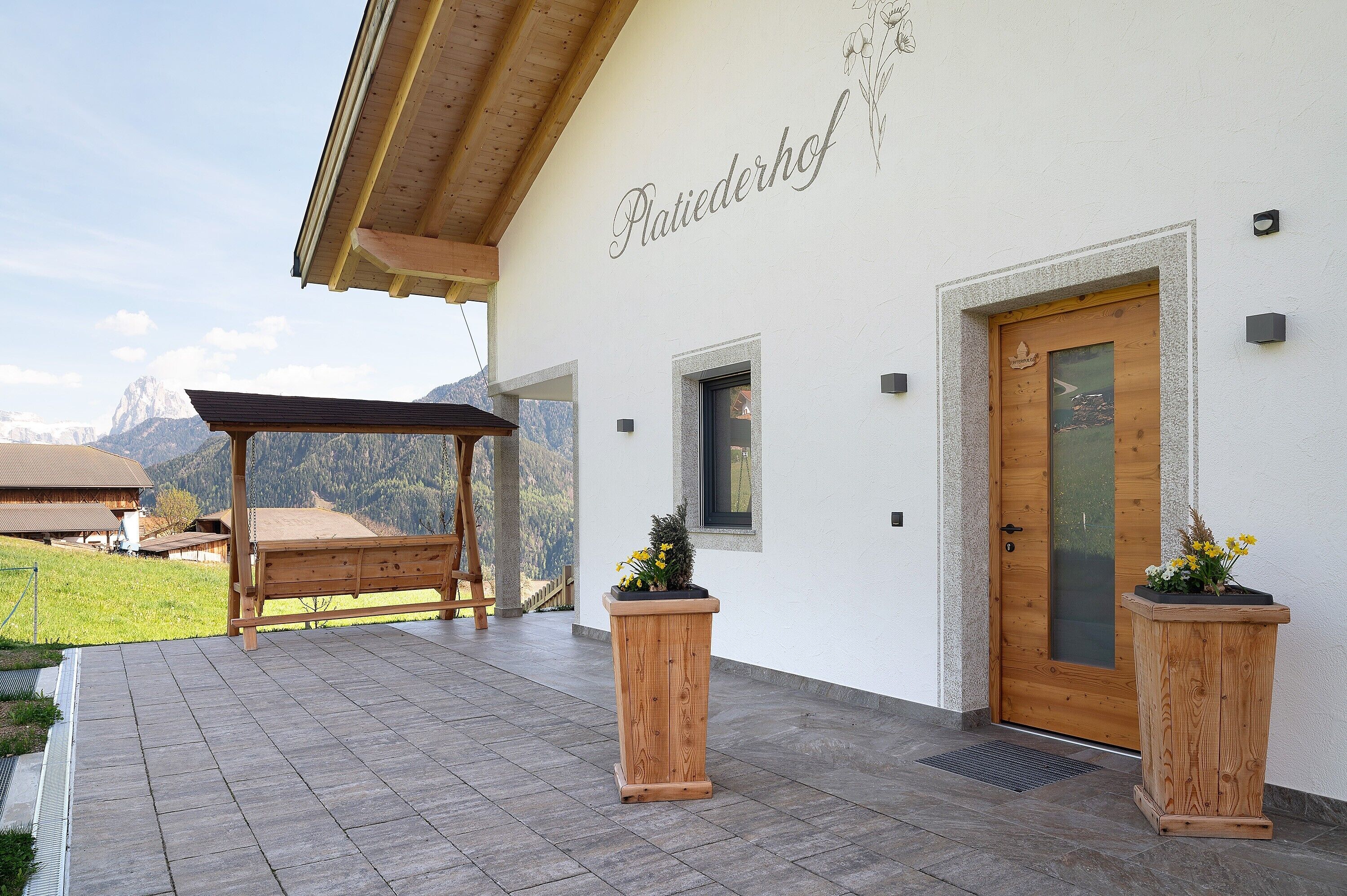 Ein Gebäude mit einer Holzpergola, einer Holztür und zwei Holzpflanzgefäßen mit Blumen.