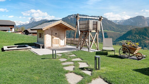 A wooden cabin with a wooden ladder, a wheelbarrow with flowers, and a stone path leading to it. - Holiday Apartment 'Platiederhof Apt Lajen' with Mountain View, Shared Garden & Wi-Fi (Laion)