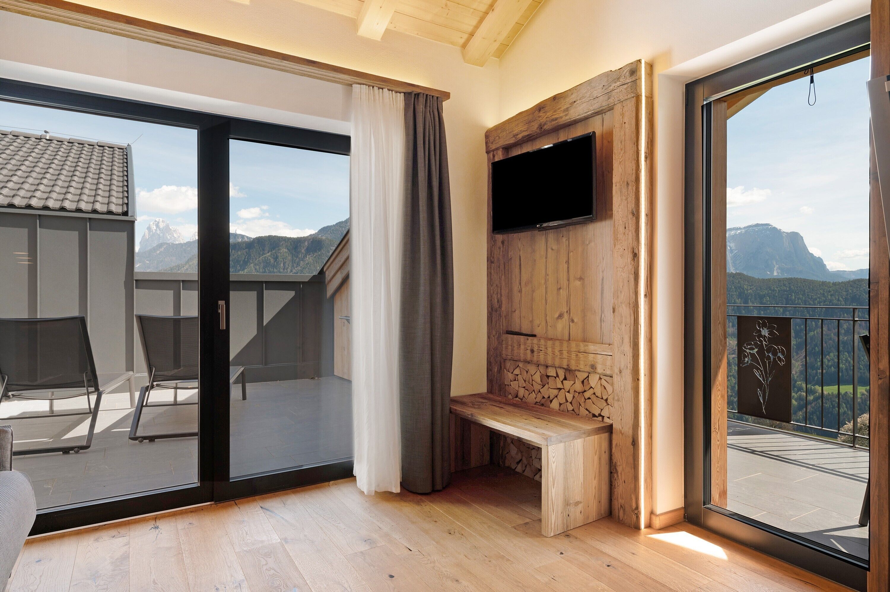 A balcony with a wooden bench, a TV mounted on a wooden wall, and a view of mountains.