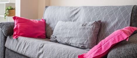 A grey sofa with patterned cushions, a wall-mounted painting, and a white shelving unit with plants.