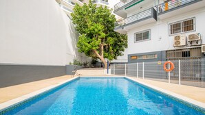 A swimming pool with a tiled edge, a tree, and a white building with balconies and air conditioning units.