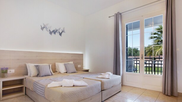 A bedroom with two beds, a wooden headboard, a nightstand, and a view of the outdoors through a sliding glass door. - JHoliday Apartment "Tropical 1" with Shared Garden, A/C & Wi-Fi (Zakynthos)