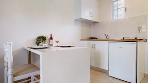 A compact kitchen with white cabinetry, a small island with a chair, a wine bottle, and a glass on the counter. - JHoliday Apartment "Tropical 1" with Shared Garden, A/C & Wi-Fi (Zakynthos)
