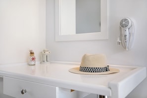 Un meuble-lavabo blanc pour la salle de bain, surmonté d’un petit objet décoratif en forme de chapeau, avec un miroir, un sèche-cheveux fixé au mur et un petit pot décoratif.