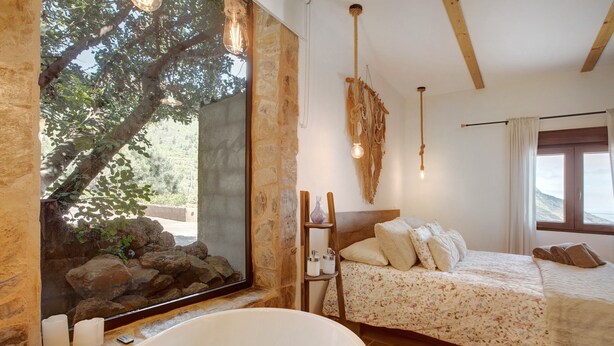 Quarto com parede de pedra, ampla janela com vista para uma árvore, piso de madeira, cama com roupa de cama floral e uma banheira.