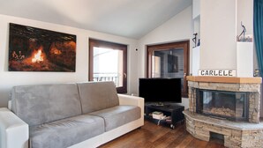 A living room with a grey sofa, a flat-screen TV, a stone fireplace with 'CARLELE' on top, and a painting of a fire on the wall. - Holiday Home "Bernhouse" with Lake View, Balcony and Wi-Fi (Dorio)