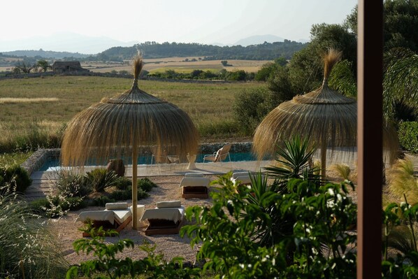 A pool area with lounge chairs and thatched umbrellas, surrounded by greenery and a view of distant hills.