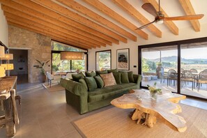 A spacious living area with a green sofa, wooden coffee table, and large windows offering a view of the outdoors. - Luxury Eco-Holiday Finca, with Stunning Panoramic Views of the Tramuntana Mountains (Sineu)