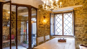 Une pièce rustique aux murs de pierre, avec une table en bois, des chaises, un lustre et une fenêtre ornée d’un motif en treillis.
