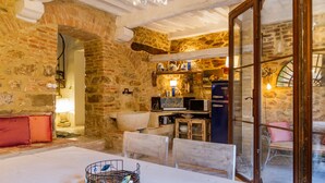 Un intérieur rustique en pierre, avec une table à manger, des chaises et un coin cuisine visible à travers une porte vitrée.