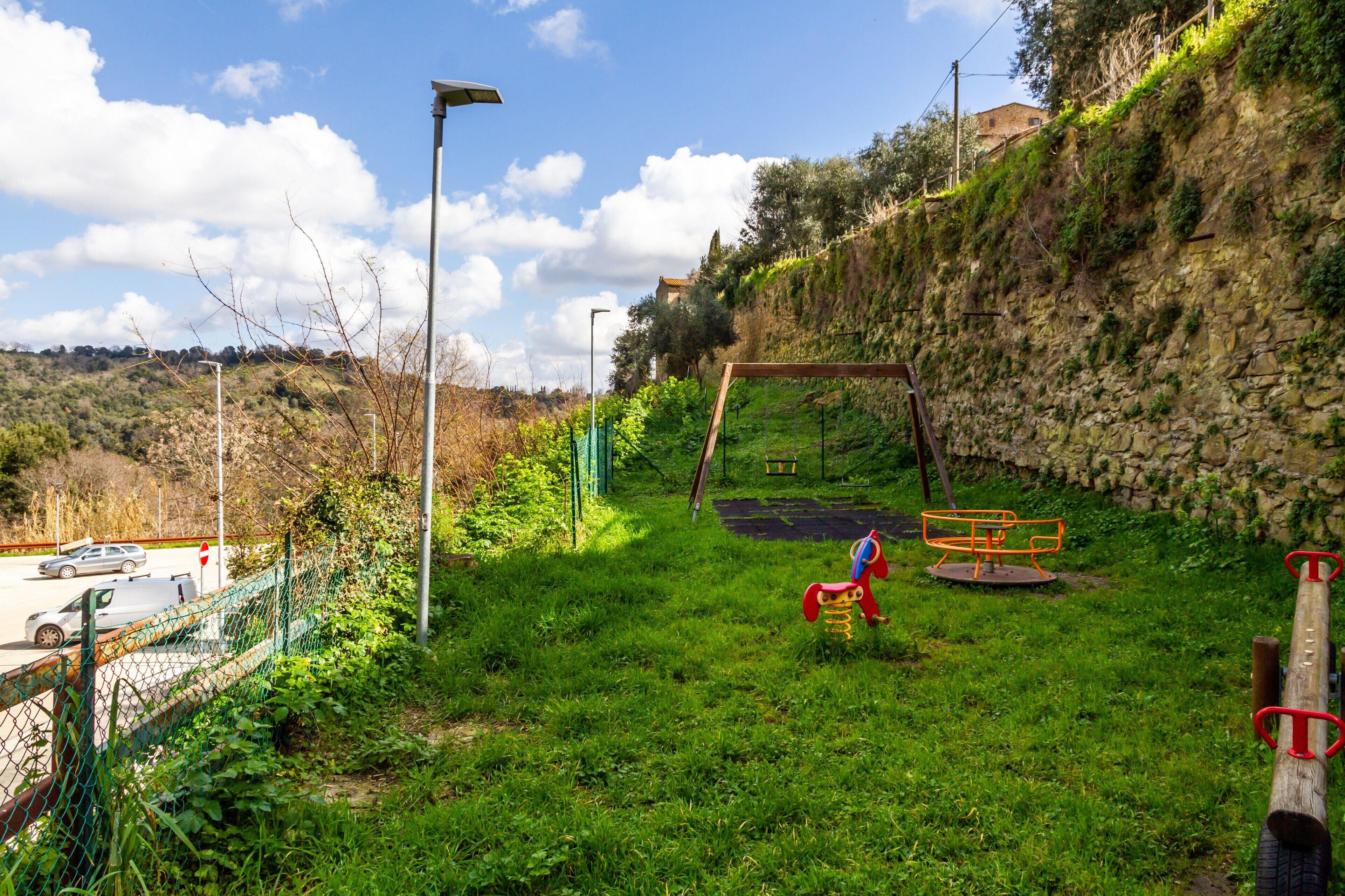 Eine Rasenfläche mit Spielplatz, einer Steinmauer und einer Straße mit Autos.