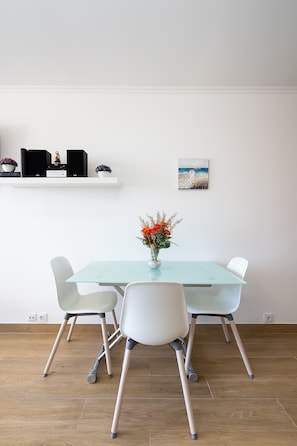 A modern dining area with a glass table, white chairs, a vase with flowers, a shelf with speakers and decorative items, and a painting on the wall.